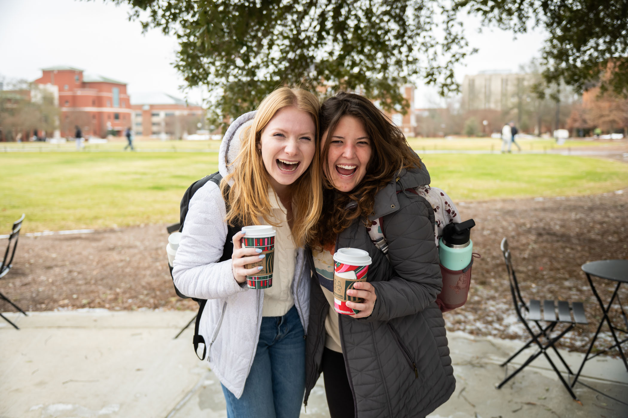 Two young women smile outdoors as they hold coffee cups